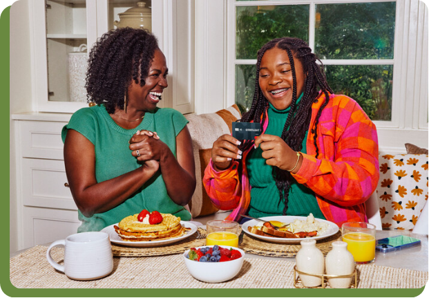Mom and daughter smiling at a Greenlight Debit Card at the breakfast table. 